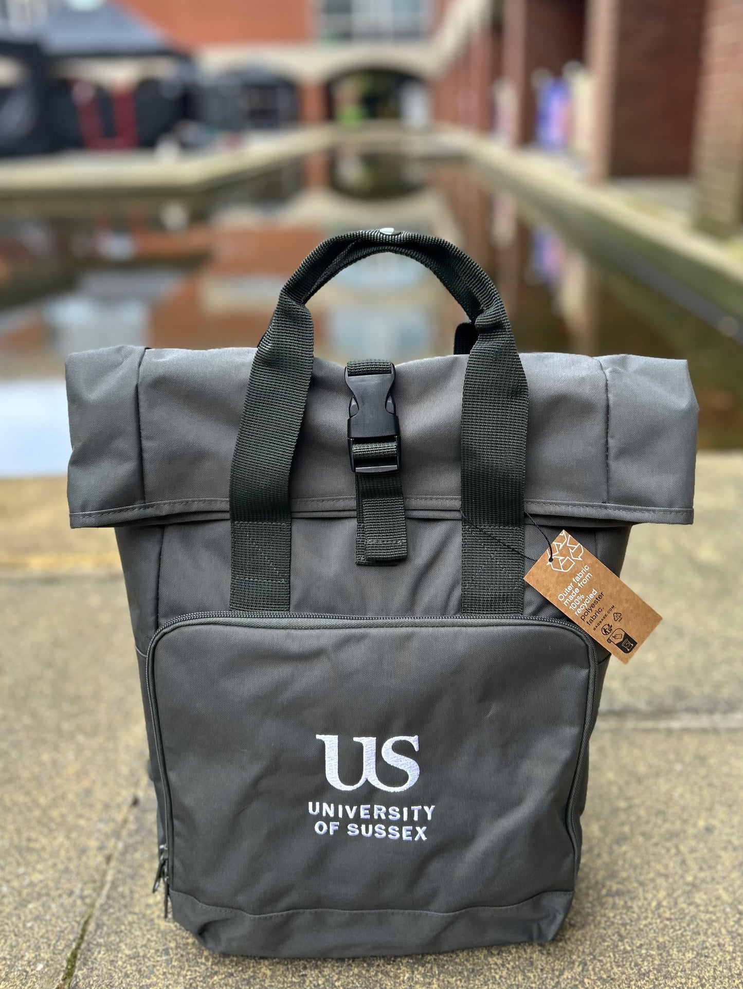 Olive green backpack with 'US University of Sussex' logo on a blurred background of the Falmer House moat and Quad