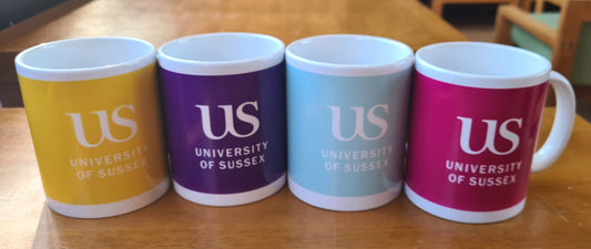 Four mugs: yellow, grape, light blue and magenta with 'US University of Sussex' branding on a wooden table in the Falmer House Common Room.