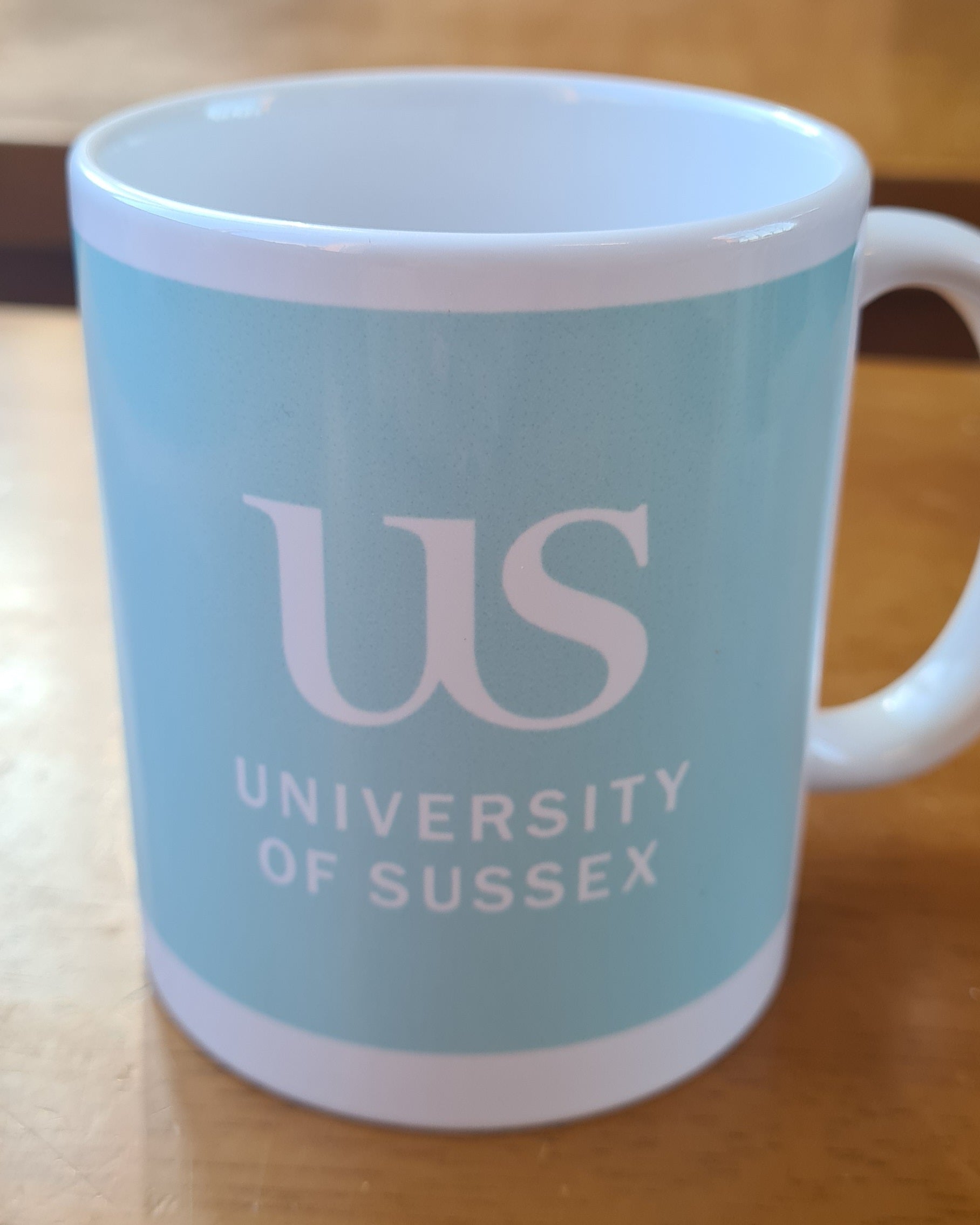 Light blue mug with the 'US University of Sussex' logo on a wooden surface
