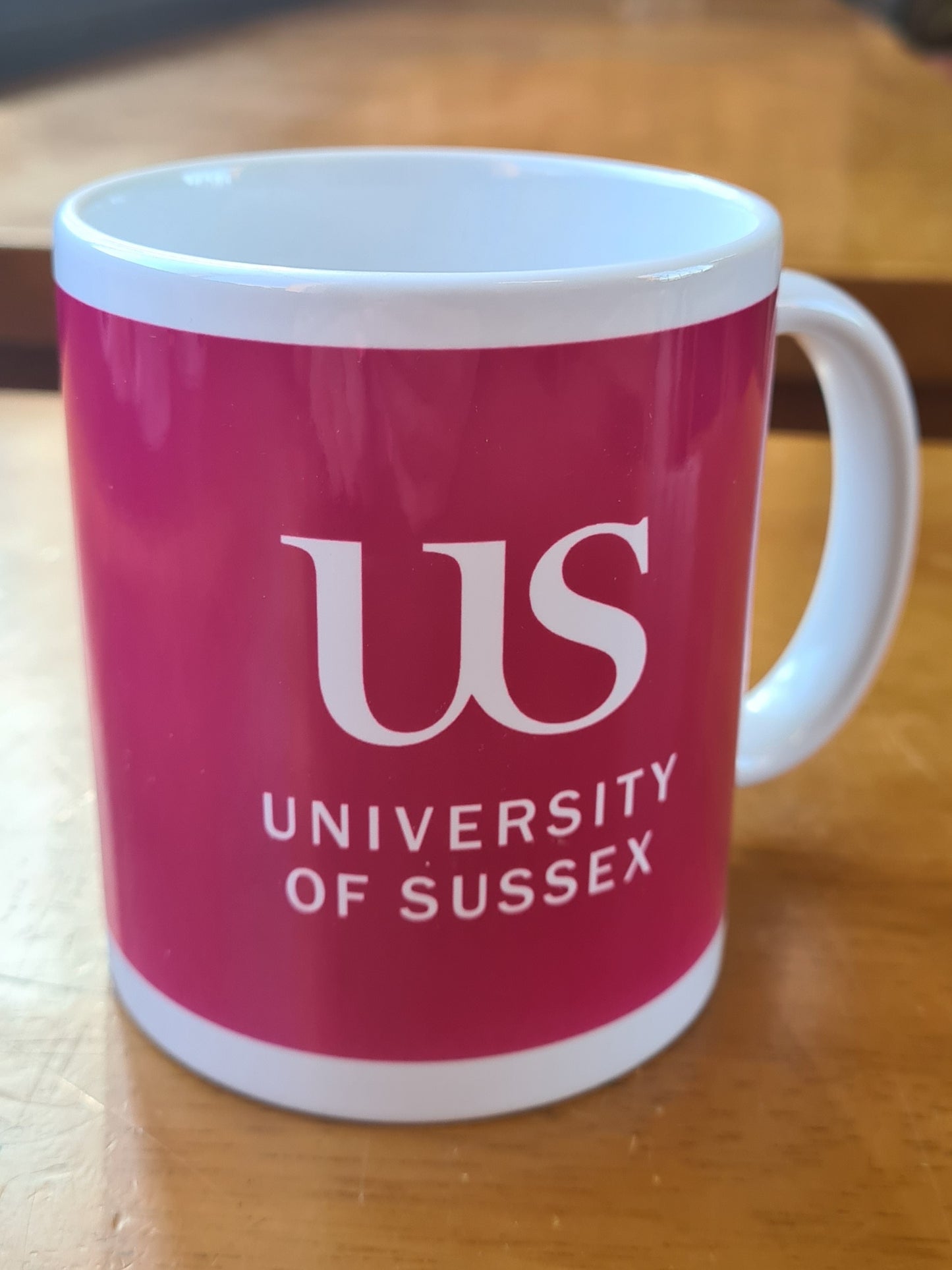 Magenta mug with the 'US University of Sussex' logo on a wooden surface