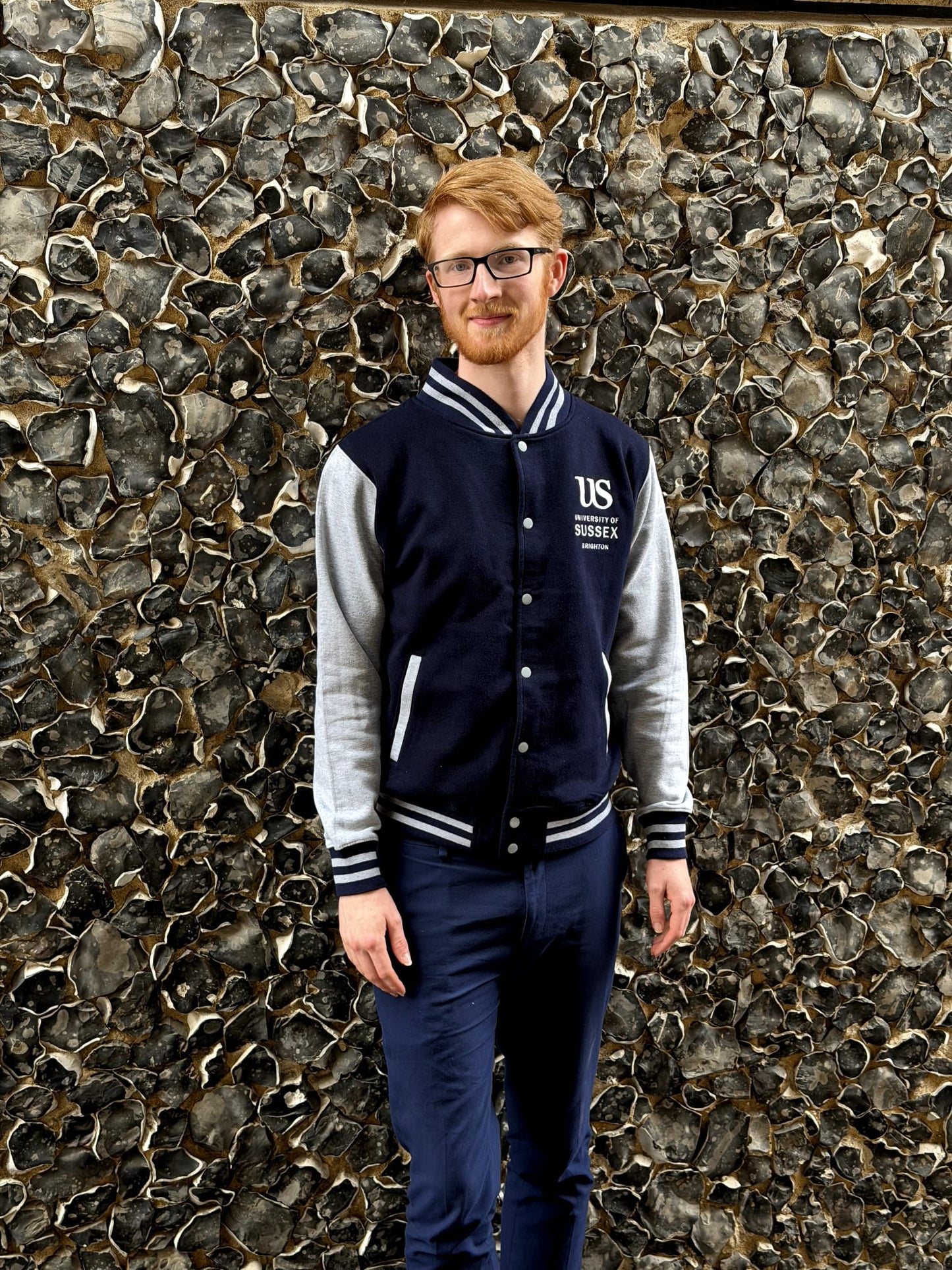 A person wearing a navy and grey Varsity jacket with the University of Sussex official logo standing in front of a textured stone wall.
