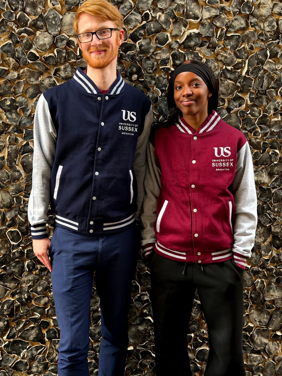 Two people, one wearing a burgundy and grey Varsity jacket and the other wearing a burgundy and grey Varsity jacket with the University of Sussex official logo standing in front of a textured stone wall.
