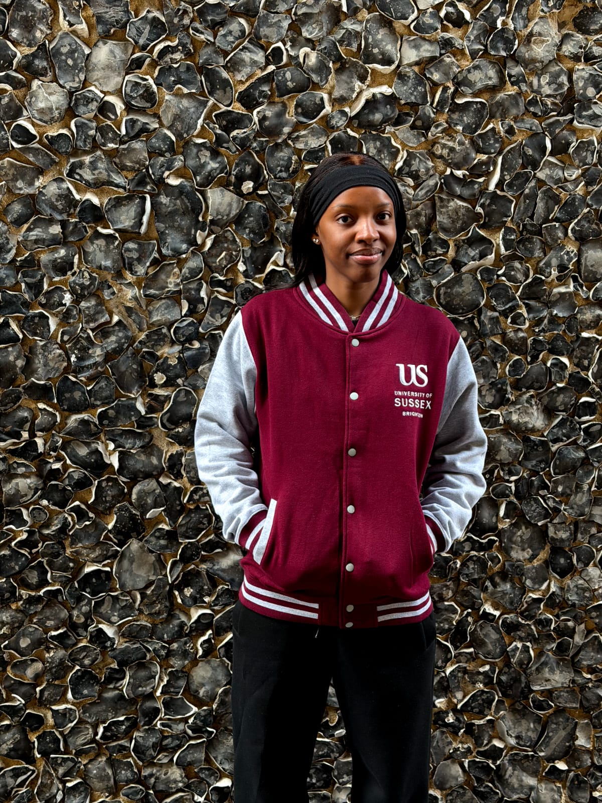 A person wearing a burgundy and gray Varsity jacket with the University of Sussex official logo standing in front of a textured stone wall.
