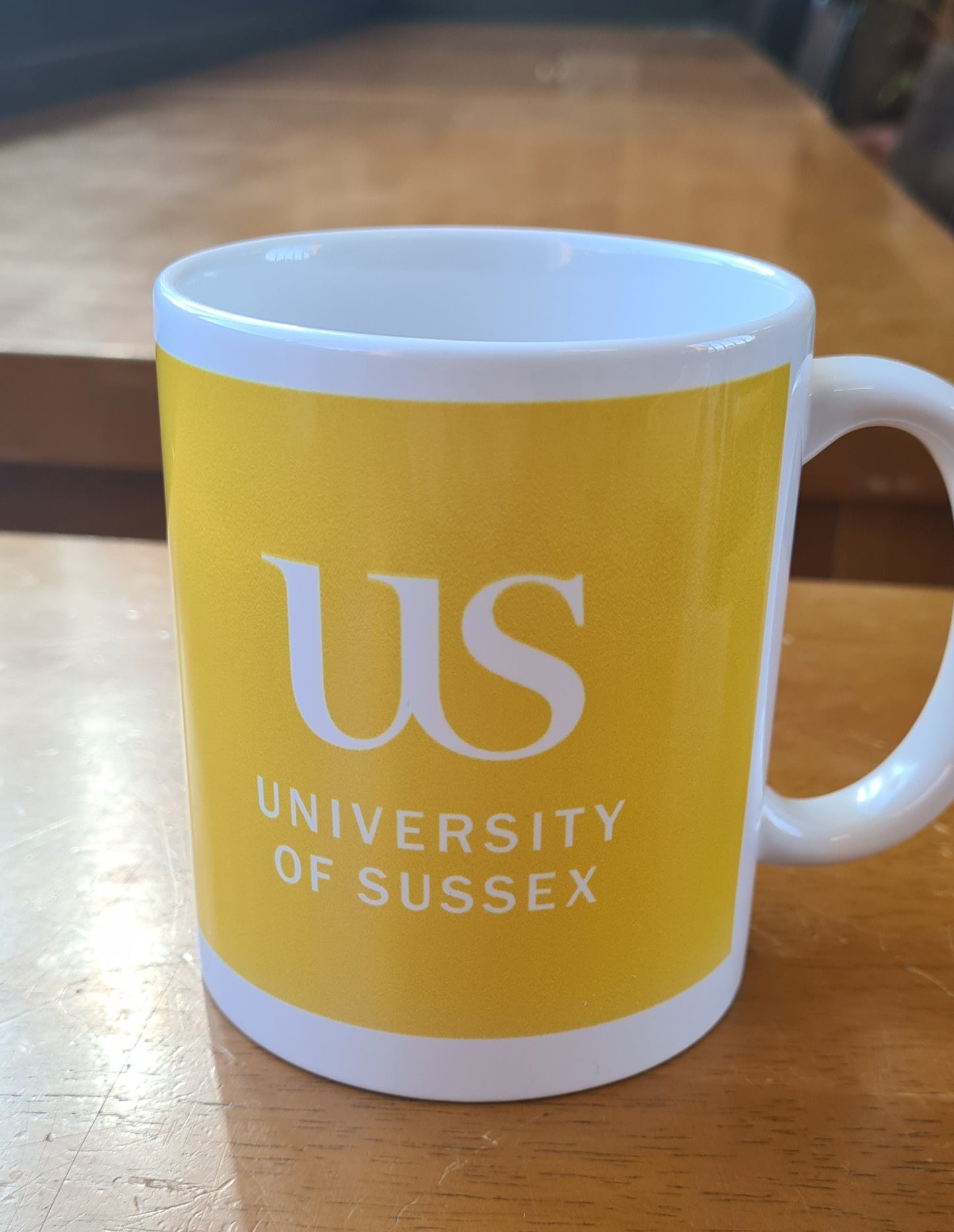 Yellow mug with the 'US University of Sussex' logo on a wooden surface